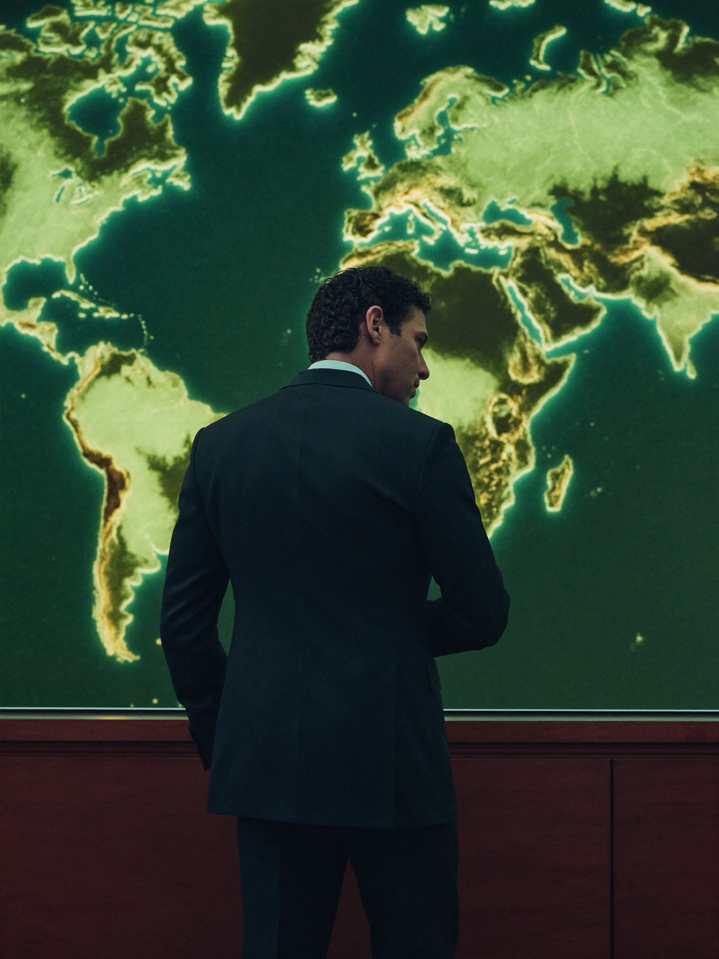 A member in a dark suit standing before the control room's glowing wall map of the world