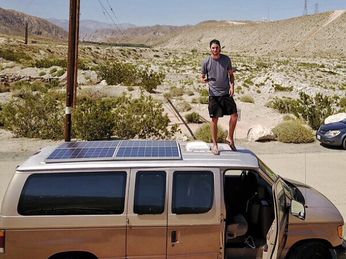 Cameron standing on the roof of the tan van he lived in, next to its solar panels, desert mountains behind.