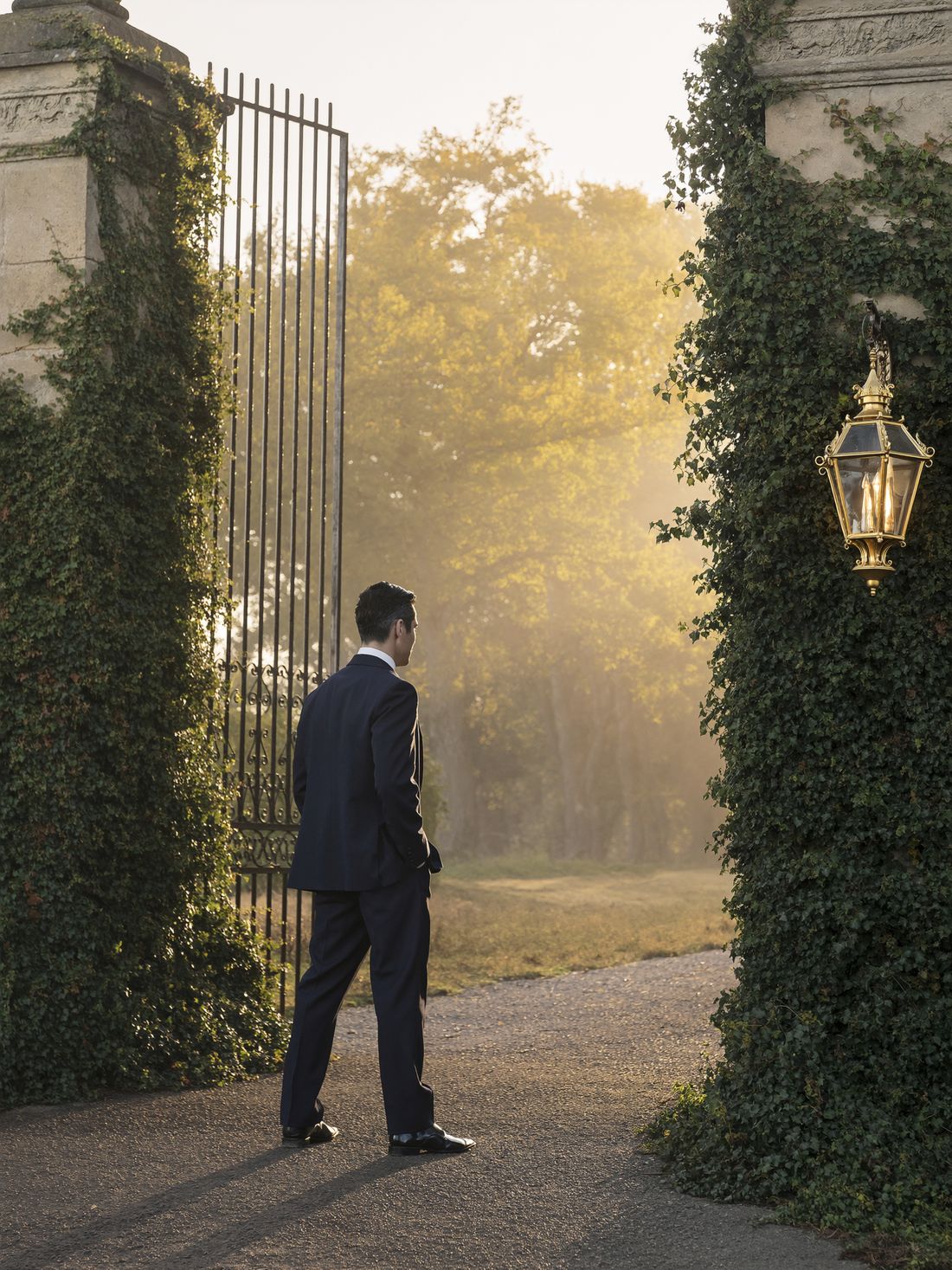 A gentleman in a navy suit standing just inside the open estate gates at dawn