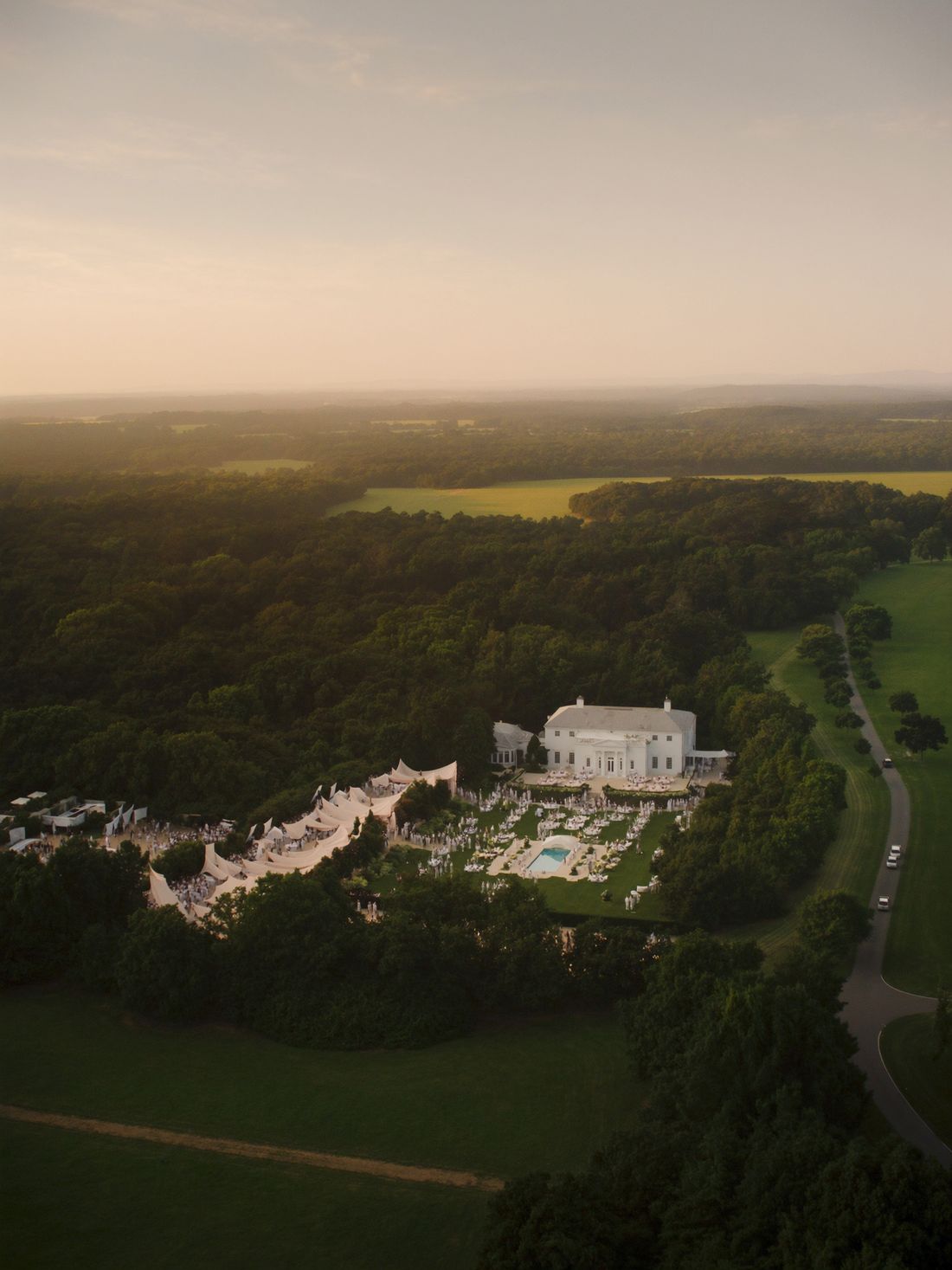 Aerial view of a white clubhouse and its grounds at dusk, tents and a pool beside the trees.