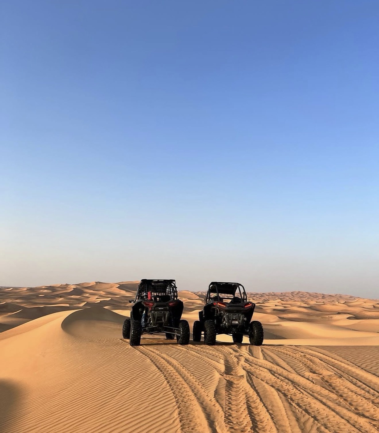 Two off-road buggies parked on the crest of a sand dune under a clear blue desert sky