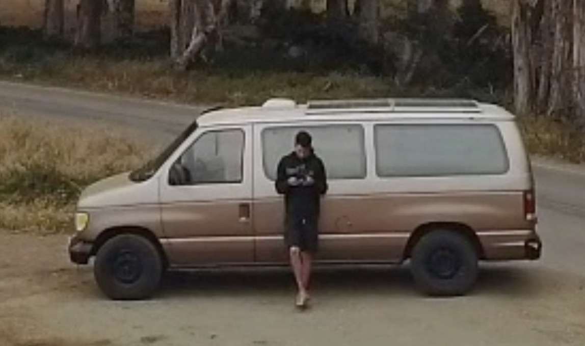 Cameron Tennant leaning against the tan van he lived in, parked on a dirt road under eucalyptus trees