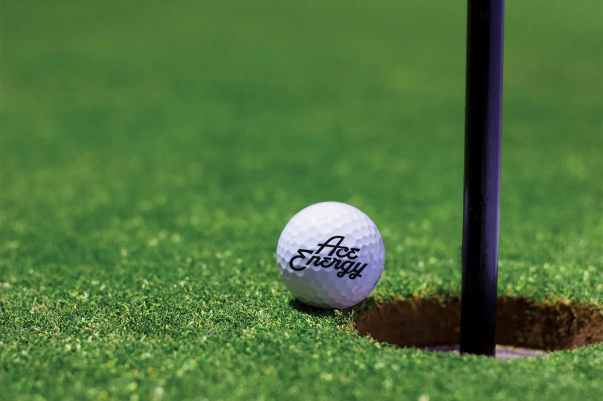 An Ace Energy branded golf ball resting at the edge of the cup on the green