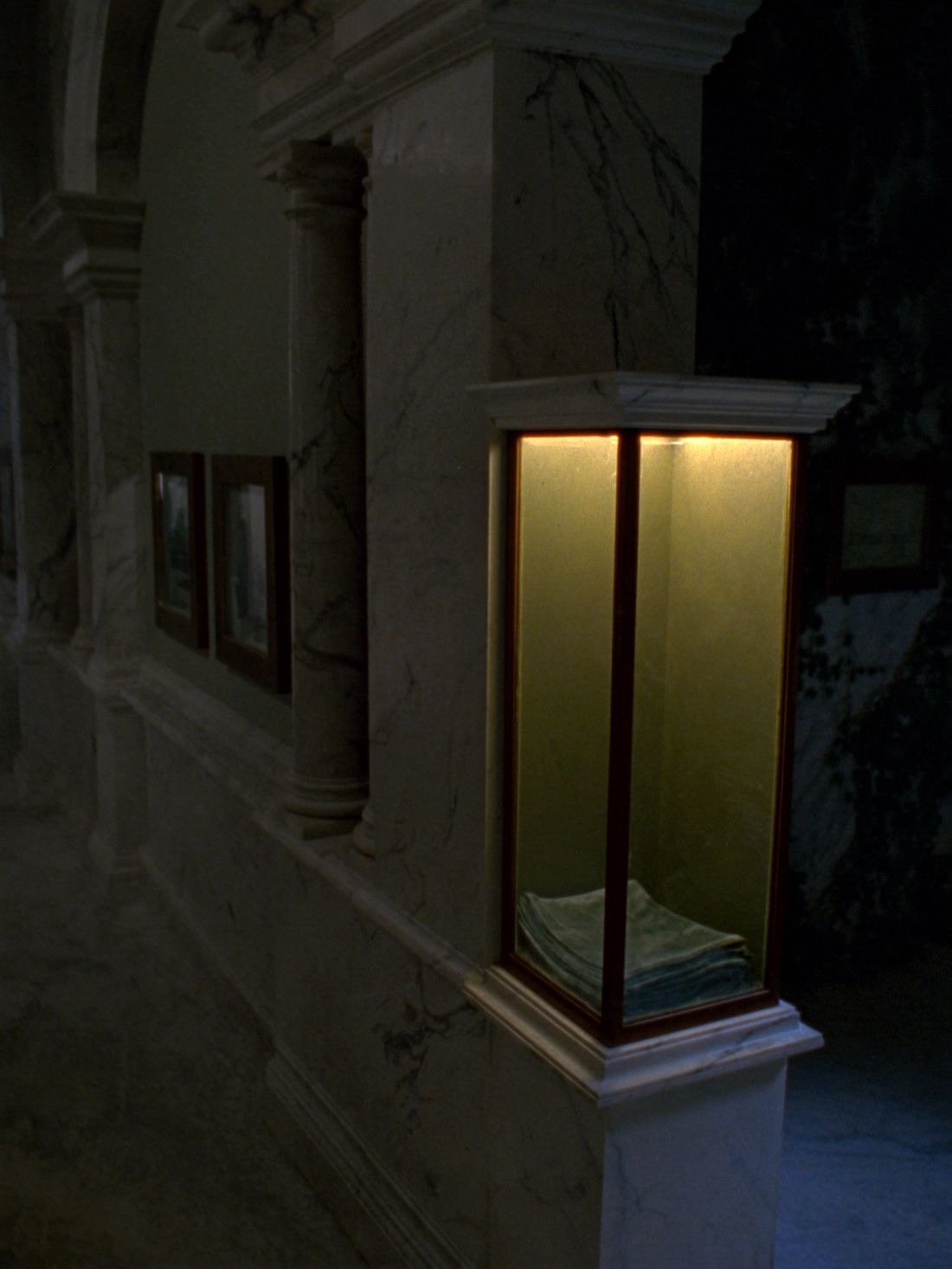 A tall empty glass display case lit gold from within, a folded green velvet cloth on its floor, marble arches behind
