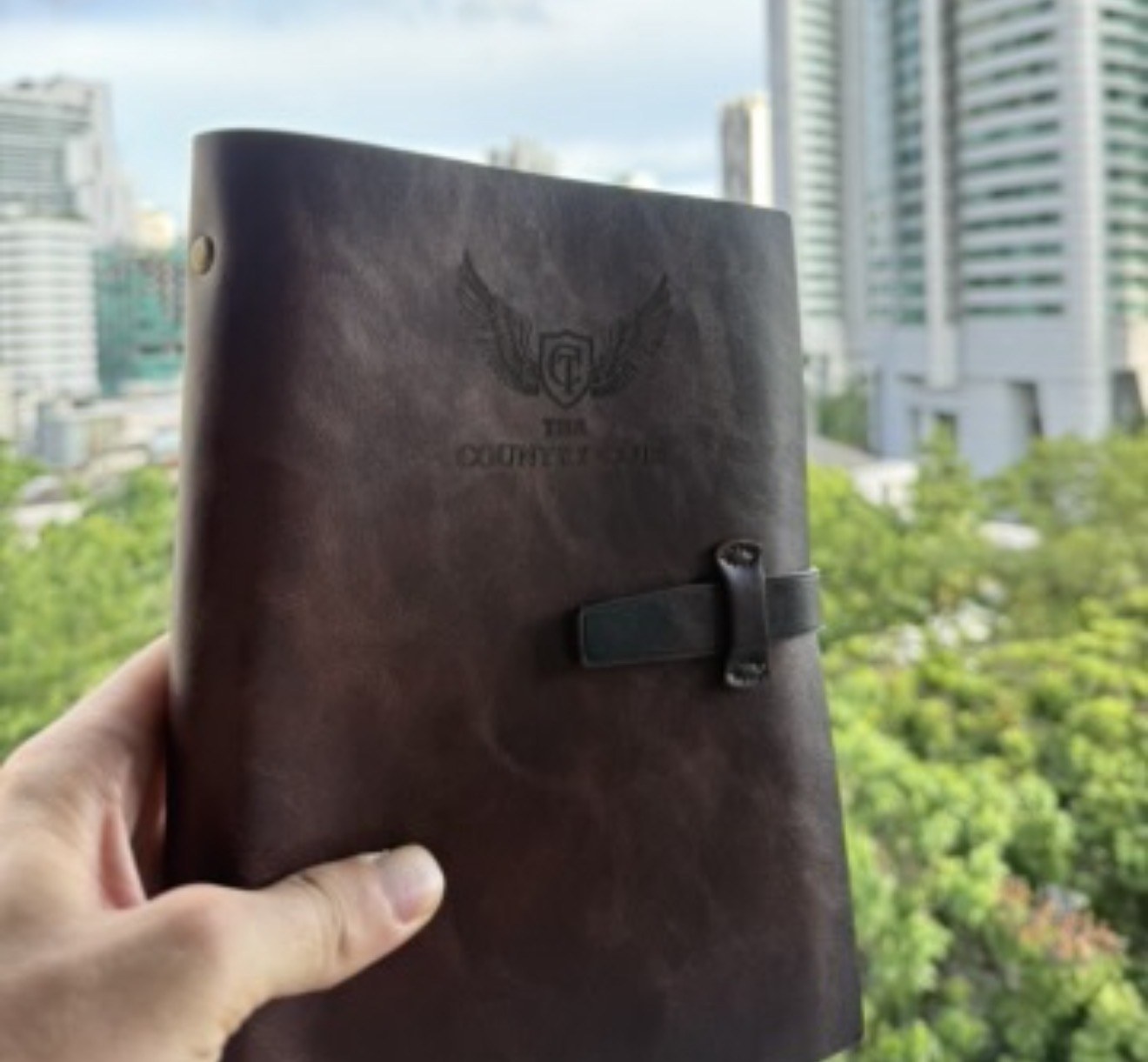 The first Country C1ub journal — a brown leather journal with the wing crest pressed into the cover and a leather strap, held up against a city skyline and green treetops