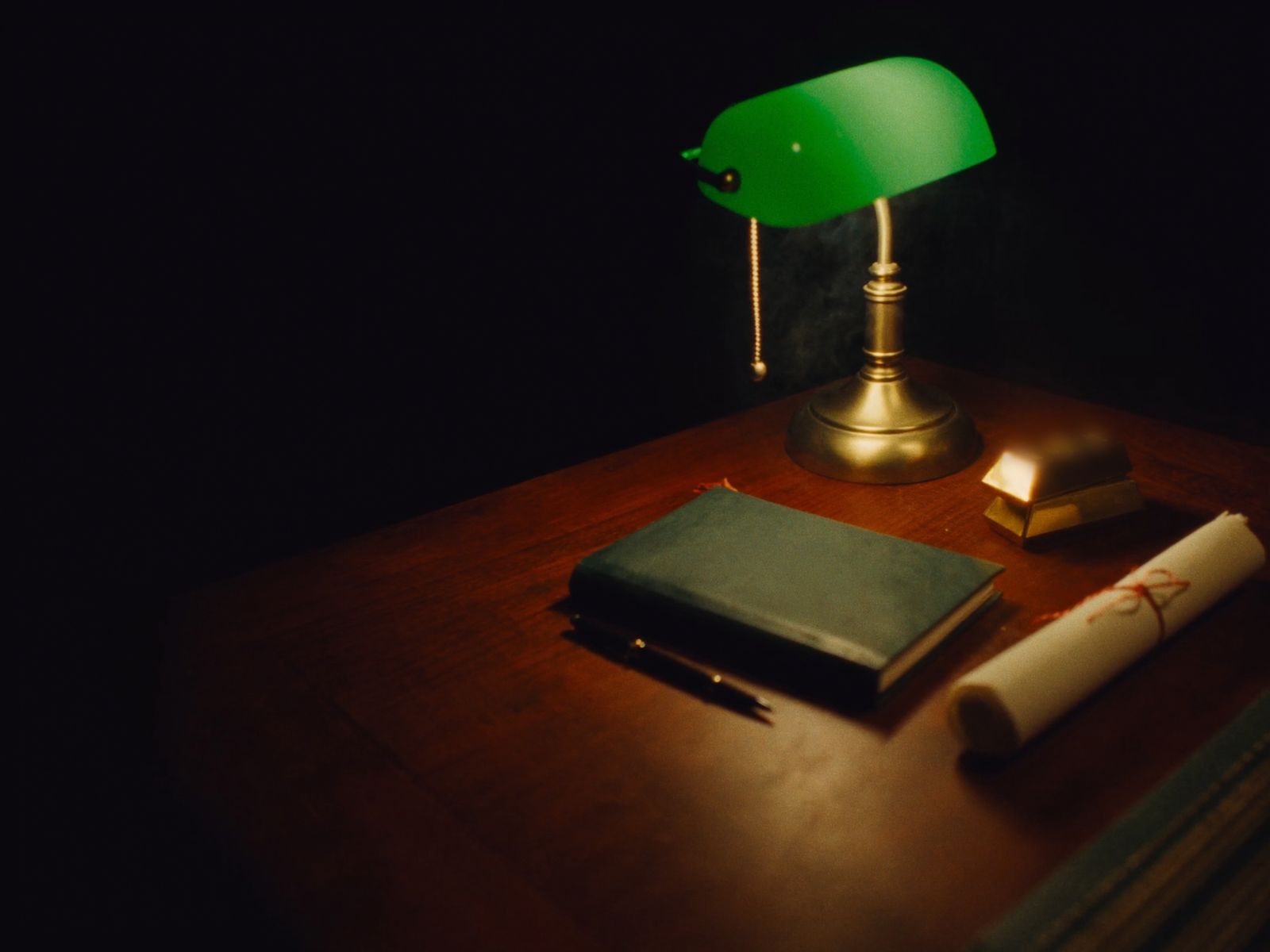A banker's lamp with a green glass shade over gold bars, a green leather ledger and a scroll tied with red string on a dark desk