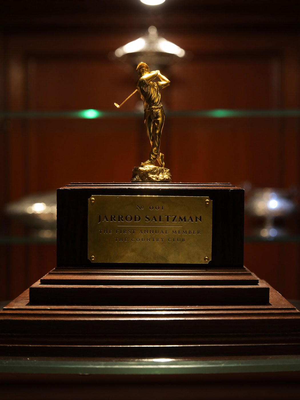 A gold statuette of a golfer at the top of his follow-through, on a dark wood plinth, inside a lit trophy cabinet — the brass plate engraved: No. 001, Jarrod Saltzman, The First Annual Member, The Country Club
