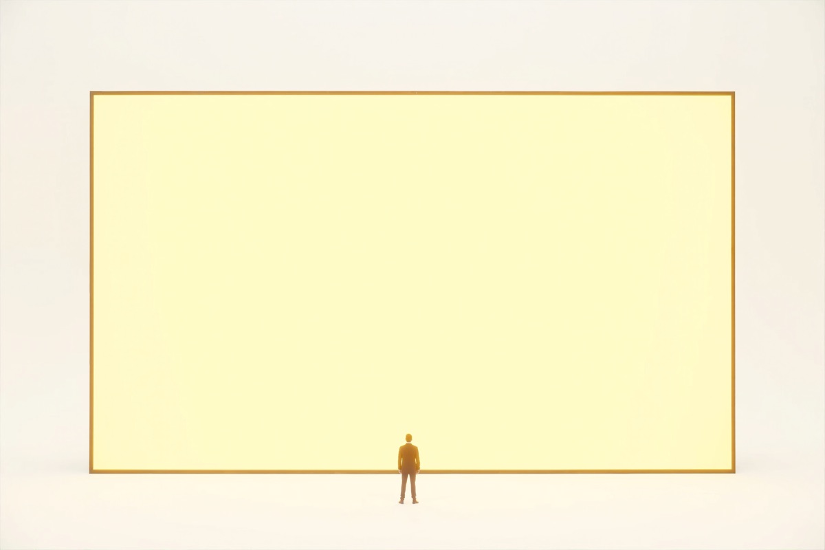 A lone figure standing small before an enormous screen glowing with warm light in the white room