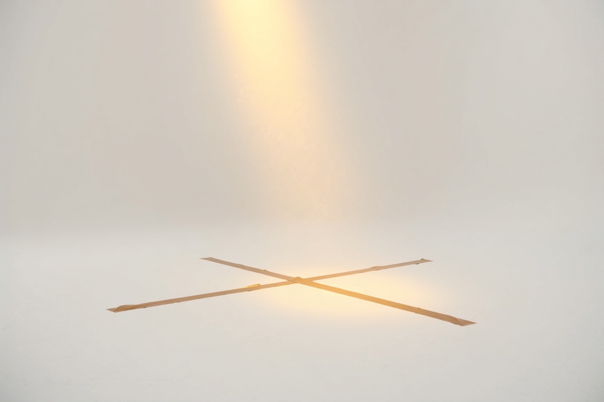 Two strips of gold tape crossed into an actor's mark on the white floor, lit by one warm spotlight